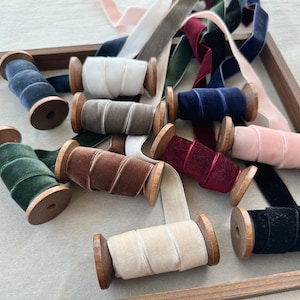 May include: Assortment of spools of velvet ribbon in various colors, including black, navy, burgundy, brown, gray, green, pink, and cream. The spools are made of wood and have a natural finish.