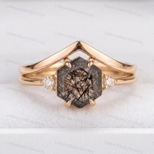Pode incluir: Um conjunto de anéis dourados. O anel central apresenta uma pedra hexagonal cinza escuro com inclusões pretas. O conjunto inclui uma banda em forma de chevrão e uma banda simples, cada uma com pequenas pedras claras. Os anéis são exibidos em um fundo claro.