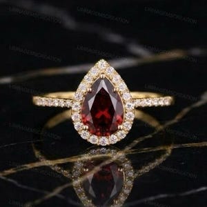 Könnte beinhalten: Ein Goldring mit einem birnenförmigen, dunkelroten Granat-Edelstein in der Mitte, umgeben von einem Kranz aus kleinen, runden, weißen Steinen. Der dünne Goldring ist zudem in der oberen Hälfte mit kleinen weißen Steinen besetzt.