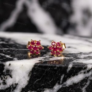 Könnte beinhalten: Ein Paar goldfarbene Ohrstecker in Blumenform. Jeder Ohrring hat eine Ansammlung von leuchtenden, runden, rubinroten Edelsteinen. Die Ohrringe werden auf einer schwarz-weißen Marmoroberfläche präsentiert.