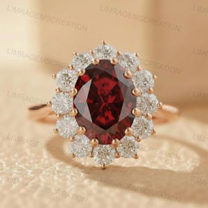 Könnte beinhalten: Ein roségoldener Ring mit einem großen, oval geschliffenen, tiefroten Edelstein, umgeben von einem Kranz aus kleineren, runden, weißen Edelsteinen. Das Design des Rings ist elegant und auffällig, perfekt für besondere Anlässe.