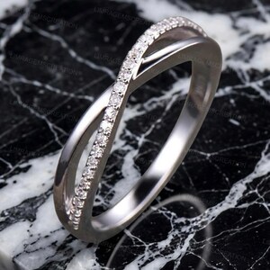 May include: A silver-colored ring with a crossover design. One band is encrusted with small, clear gemstones. The ring is set against a black and white marble-patterned surface. The ring's design is modern and elegant.
