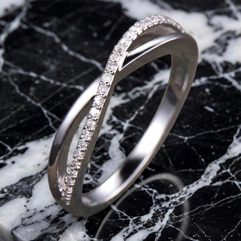 May include: A silver-colored ring with a crossover design. One band is encrusted with small, clear gemstones. The ring is set against a black and white marble-patterned surface. The ring's design is modern and elegant.