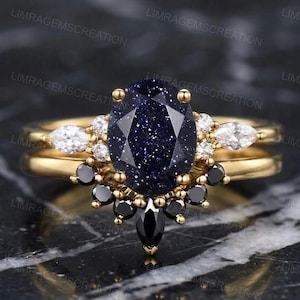 Könnte beinhalten: Ein goldener Ringsatz mit einem großen, ovalen, dunkelblauen Edelstein mit funkelnden Einschlüssen. Der Ring ist mit weißen und schwarzen Edelsteinen akzentuiert. Das Band hat eine einfache goldene Farbe. Der Ring ist auf einer dunklen, strukturierten Oberfläche platziert.