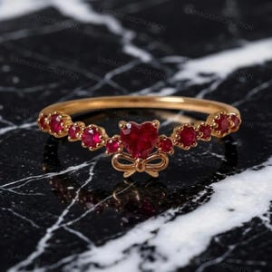 Könnte beinhalten: Ein goldener Ring mit einem zentralen herzförmigen roten Edelstein, der von kleineren runden roten Edelsteinen entlang der Ringschiene flankiert wird. Ein kleines goldenes Schleifendetail sitzt direkt unter dem herzförmigen Stein.