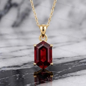 Könnte beinhalten: Eine goldfarbene Halskette mit einem Anhänger, der einen großen, rechteckigen, tiefroten Edelstein aufweist. Der Edelstein ist facettiert und in eine goldene Fassung gesetzt. Die Kette ist filigran und ebenfalls goldfarben.
