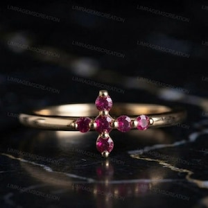 Könnte beinhalten: Ein goldener Ring mit einer kreuzförmigen Anordnung aus fünf runden, leuchtend rosaroten Edelsteinen. Das Ringband ist dünn und poliert und zeigt ein schlichtes Design, bei dem die Steine direkt in die Mitte der Ringoberseite eingefasst sind.
