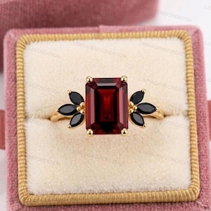 Puede incluir: Un anillo de oro con una gran piedra preciosa rectangular de color rojo oscuro en el centro. A ambos lados de la piedra central hay grupos de piedras negras en forma de lágrima. El anillo se presenta sobre un fondo de terciopelo rosa.
