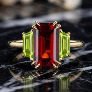 Könnte beinhalten: Ein goldener Ring mit einem großen, rechteckigen, tiefroten Edelstein, flankiert von zwei kleineren, rechteckigen, hellgrünen Edelsteinen. Die Fassung des Rings hat ein strukturiertes, klauenartiges Design. Der Ring wird auf einer dunklen, reflektierenden Oberfläche präsentiert.