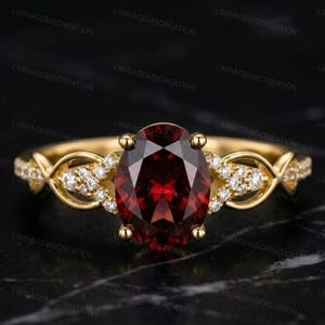 Könnte beinhalten: Ein Goldring mit einem großen, oval geschliffenen, tiefroten Granat in der Mitte. Die Ringschiene ist auf beiden Seiten mit einem komplexen, durchbrochenen Design gestaltet, das mit kleinen, klaren, rund geschliffenen weißen Steinen besetzt ist.
