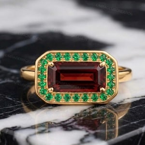 Könnte beinhalten: Ein Goldring mit einem rechteckigen, im Smaragdschliff gehaltenen, tiefroten Granat als Mittelstein. Der Granat ist von einem Kranz aus kleinen, runden, leuchtend grünen Smaragden in einer Goldfassung umgeben. Die Ringschiene ist aus glattem Gold.