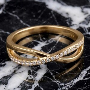May include: A gold-colored ring with a crisscross design. One band is encrusted with small, clear gemstones. The ring is set against a black and white marble surface. The ring is marked with "14K".