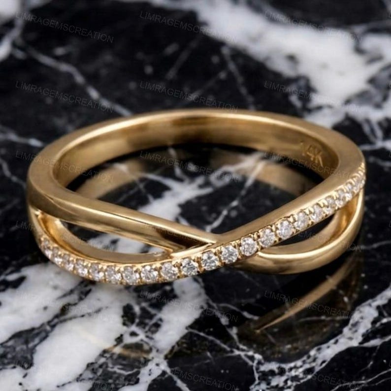 May include: A gold-colored ring with a crisscross design. One band is encrusted with small, clear gemstones. The ring is set against a black and white marble surface. The ring is marked with "14K".