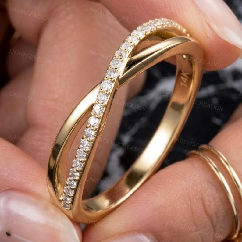 May include: A gold-colored ring with a crisscross design. One band is encrusted with small, clear gemstones. The ring is marked with "14K". The ring is held by a person.