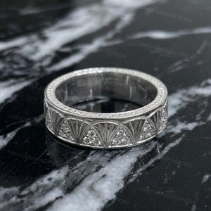 Könnte beinhalten: Ein silberner Ring mit einem detaillierten geometrischen Design. Der Ring zeigt ein sich wiederholendes Muster aus fächerförmigen Elementen, die jeweils mit kleinen, funkelnden Steinen verziert sind. Das Band ist ebenfalls mit kleinen Steinen besetzt, was den Glanz des Rings verstärkt.