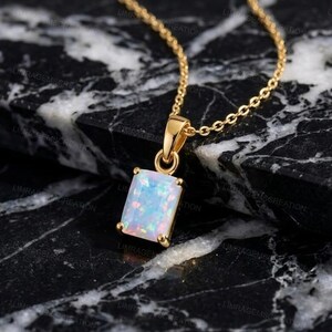 Könnte beinhalten: Eine Goldhalskette mit einem rechteckigen Anhänger, der einen weißen Opal mit irisierenden Farbreflexen enthält. Der Anhänger ist an einer goldenen Öse befestigt und mit einer feinen Goldkette verbunden, die ein poliertes Metallfinish aufweist.