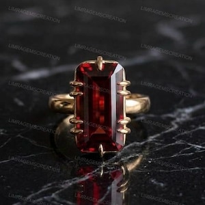 Könnte beinhalten: Ein Goldring mit einem großen, rechteckigen, tiefroten Edelstein, der vertikal gefasst ist. Der Stein wird von sechs markanten, krallenartigen Metallfassungen auf jeder Seite gehalten, was ein strukturiertes, symmetrisches Design ergibt.
