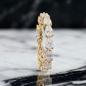 Könnte beinhalten: Ein goldener Ring mit einer durchgehenden Reihe von birnenförmigen Diamanten. Das Ringband ist ebenfalls goldfarben und weist zwischen den Diamanten ein filigranes, offenes Design auf. Die Diamanten funkeln vor einem Marmorhintergrund.