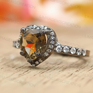 Könnte beinhalten: Ein herzförmiger Ring mit einem großen, facettierten, bernsteinfarbenen Edelstein. Der Edelstein ist von kleineren, klaren Steinen umgeben und in einem dunklen Metallband gefasst. Der Ring wird auf einer hellen Holzoberfläche präsentiert.
