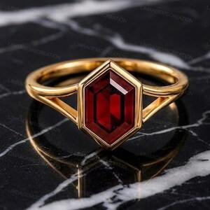 Könnte beinhalten: Ein Goldring mit einem zentralen, sechseckig geschliffenen, tiefroten Edelstein in einer Zargenfassung. Die Ringschiene hat ein geteiltes Design, das sich zum Mittelstein hin verjüngt. Das Metall hat eine polierte Oberfläche mit Glanz.
