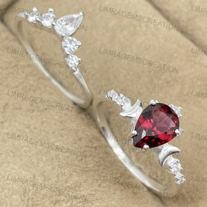 Könnte beinhalten: Zwei Silberringe. Ein Ring hat einen birnenförmigen, tiefroten Edelstein. Der andere Ring hat eine Reihe von klaren, runden und birnenförmigen Edelsteinen. Beide Ringe haben kleine, klare Akzentsteine.