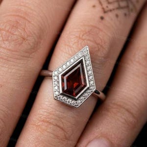 Könnte beinhalten: Ein silberfarbener Ring mit einem drachenförmigen, tiefroten Edelstein in der Mitte. Der zentrale Stein ist von einem Kranz aus kleinen, klaren, rund geschliffenen Steinen umgeben, die in den Metallrahmen gefasst sind, der in eine dünne Ringschiene übergeht.