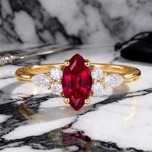 May include: A gold ring featuring a large, marquise-cut ruby center stone, flanked by clusters of smaller, round, and pear-shaped white gemstones. The ring has a polished gold band and a classic, elegant design.