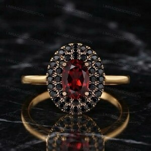 Könnte beinhalten: Ein Goldring mit einem zentralen, oval geschliffenen, tiefroten Granat-Edelstein, der von einem Kranz aus kleinen, runden schwarzen Steinen umgeben ist. Die Ringschiene ist aus glattem, poliertem Gold und bildet einen Kontrast.