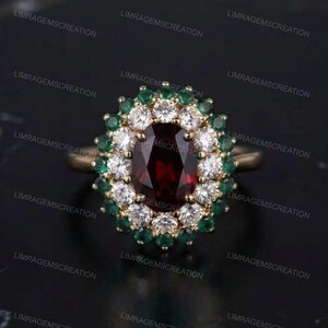 Könnte beinhalten: Ein Goldring mit einem zentralen, oval geschliffenen, tiefroten Granat-Edelstein. Der Granat ist von einem Kranz aus runden weißen Diamanten umgeben, der wiederum von einem zweiten Kranz aus kleinen, runden grünen Smaragden in Gold gefasst ist.