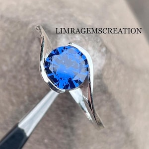 Könnte beinhalten: Ein silberner Ring mit einem großen, runden, brillantgeschliffenen blauen Edelstein. Das Band des Rings wölbt sich nach oben, um den Edelstein zu halten. Der Text "LIMRAGEMSCREATION" ist sichtbar.