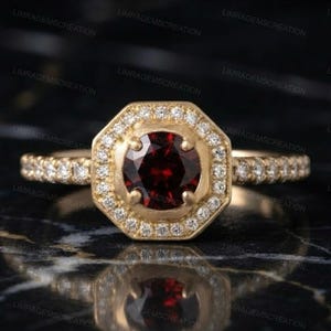 Könnte beinhalten: Ein goldener Ring mit einem runden, tiefroten Mittelstein, der in einem achteckigen Heiligenschein aus kleinen, klaren, funkelnden Steinen gefasst ist. Das Band ist zudem mit einer Reihe kleiner, klarer Steine an jeder Seite verziert.