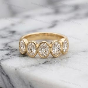 Könnte beinhalten: Ein goldener Bandring mit fünf oval geschliffenen, klaren Steinen in einer Reihe. Jeder Stein ist in einer glatten Zargenfassung gefasst, wodurch eine durchgehende Linie facettierter Edelsteine auf der Vorderseite des Rings entsteht. Das Metall ist poliert.