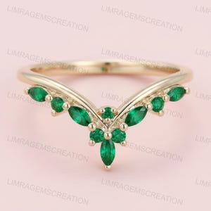 Könnte beinhalten: Ein goldfarbener Ring mit V-förmigem Design. Der Ring ist mit mehreren smaragdgrünen Edelsteinen besetzt. Die Edelsteine sind in einem floralen Muster in der Mitte und entlang der Kurve des Rings angeordnet, was einen zarten und eleganten Look erzeugt.