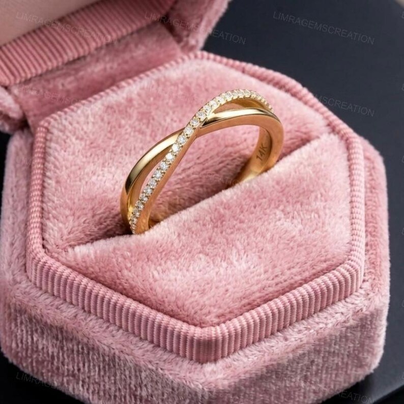 May include: A gold-colored ring with a crisscross design, encrusted with small, clear gemstones. The ring is set in a pink velvet jewelry box. The ring is marked with "14K".
