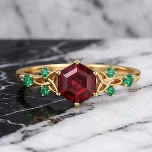 Könnte beinhalten: Ein goldener Ring mit einem großen, sechseckigen, tiefroten Edelstein im Zentrum. Der Ring ist mit kleineren grünen Edelsteinen und dekorativen goldenen Akzenten am Band verziert. Der Ring ist vor einem Marmorhintergrund platziert.