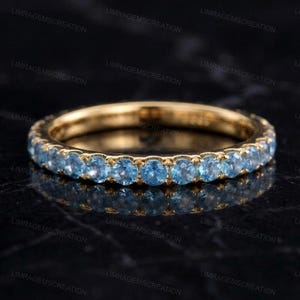 May include: A gold band ring set with a row of round, light blue gemstones. The ring is displayed on a dark, reflective surface, highlighting the sparkle of the gemstones. The ring is a piece of jewelry.