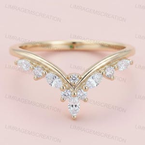 Könnte beinhalten: Ein goldfarbener Ring mit geschwungenem Design, besetzt mit mehreren klaren, funkelnden Edelsteinen. Der Ring hat ein V-förmiges Band und eine Ansammlung von Steinen in der Mitte, was ihm ein zartes und elegantes Aussehen verleiht. Der Ring ist vor einem hellrosa Hintergrund.