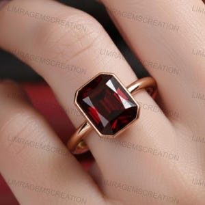Könnte beinhalten: Ein roségoldener Ring mit einem rechteckigen, tiefroten Edelstein. Der Edelstein ist in einer Zargenfassung gefasst, und der Ring ist schlicht und poliert. Der Ring wird an einem Finger getragen.