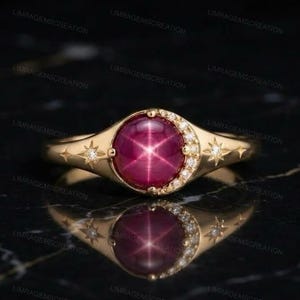 Könnte beinhalten: Ein Goldring mit einem runden, tiefrosa Stern-Edelstein im Cabochon-Schliff. Die Ringschiene ist mit einer Mondsichel aus kleinen klaren Steinen sowie zwei sternförmigen Gravuren verziert, die jeweils einen winzigen klaren Stein enthalten.