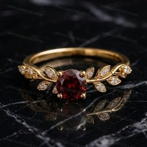Könnte beinhalten: Ein goldener Ring mit einem runden, tiefroten Granat-Edelstein in der Mitte. Das Band ist mit einer Reihe kleiner, metallischer blattförmiger Motive gestaltet, die vom Zentrum ausgehen und jeweils mit winzigen, klaren, weißen Steinen besetzt sind.