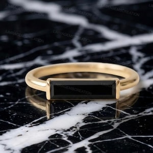 Könnte beinhalten: Ein goldener Ring mit einem horizontalen, rechteckigen schwarzen Edelstein, der in ein glattes, poliertes Goldband eingefasst ist. Der Ring hat ein klares, lineares Design mit dem dunklen Stein in der Mitte, was einen starken Kontrast zwischen Metall und Stein erzeugt.