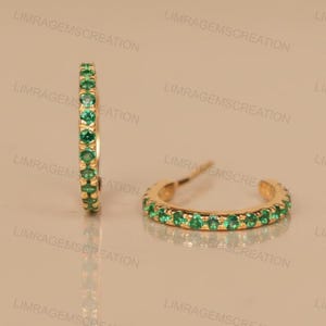 May include: Gold hoop earrings with a row of small, vibrant green gemstones. The earrings are set in a gold metal and have a polished finish. The design is simple and elegant, suitable for everyday wear.