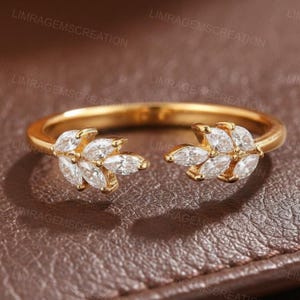 Könnte beinhalten: Ein goldfarbener Ring mit geteiltem Schaftdesign. Der Ring hat zwei blattartige Cluster, die jeweils aus marquiseförmigen, klaren Edelsteinen bestehen. Der Ring ist vor einem braunen Hintergrund platziert.