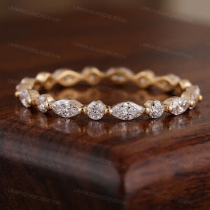 Könnte beinhalten: Ein goldfarbener Ring mit abwechselnd runden und marquiseförmigen, klaren Edelsteinen. Der Ring liegt auf einer dunklen Holzoberfläche. Ein filigranes Schmuckstück für jeden Anlass.
