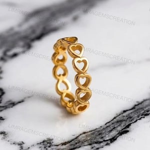 Könnte beinhalten: Ein goldfarbener Ring mit einem sich wiederholenden Herzmuster. Der Ring besteht aus poliertem Metall und liegt auf einer Marmoroberfläche. Das Design besteht aus mehreren Herzen, die zu einem durchgehenden Band verbunden sind. Ein schönes Schmuckstück.