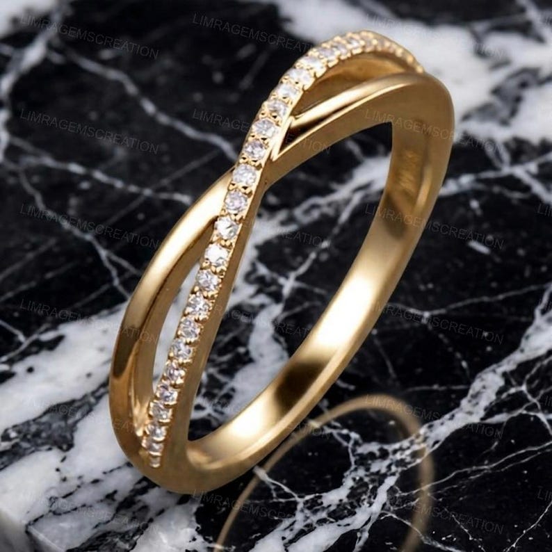 May include: A gold-colored ring with a crisscross design. One band is encrusted with small, clear gemstones. The ring is set against a black and white marble surface, creating a contrast that highlights the ring's details.