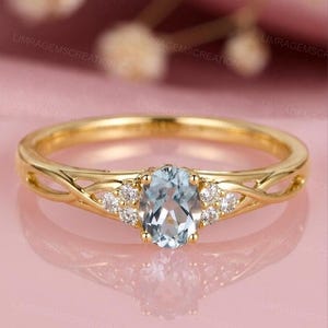 Op de afbeelding: Een gouden ring met een ovale, lichtblauwe edelsteen in het midden, geflankeerd door kleinere witte edelstenen. De band heeft een ingewikkeld, verweven ontwerp. De ring is geplaatst tegen een zachte roze achtergrond.
