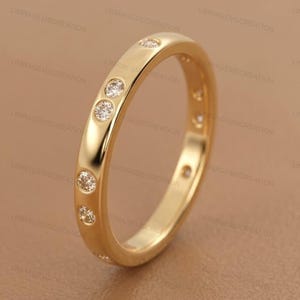 Könnte beinhalten: Ein goldfarbener Ring mit kleinen, klaren Edelsteinen, die um das Band herum eingesetzt sind. Der Ring hat eine glatte, polierte Oberfläche und wird vor einem neutralen Hintergrund gezeigt. Der Ring ist ein Schmuckstück.