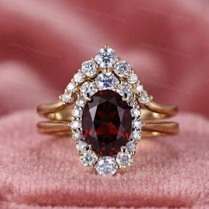 Könnte beinhalten: Ein Goldring-Set mit einem zentralen, oval geschliffenen, tiefroten Granat, der von einem Kranz aus kleinen, runden weißen Steinen umgeben ist. Das Design umfasst einen passenden, geschwungenen Ring, der ebenfalls mit weißen Steinen besetzt ist.
