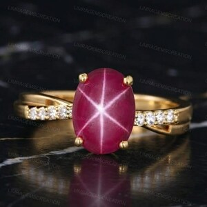 Könnte beinhalten: Ein Goldring mit einem zentralen, ovalen Sternrubin-Cabochon, der ein markantes sechsstrahliges Sternmuster aufweist. Die Ringschiene ist aus Gelbgold gefertigt und mit kleinen, klaren, rund geschliffenen Steinen entlang der Seiten besetzt.